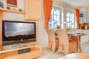 Eine moderne Küche mit weißen Schränken, eine Essecke mit Korbstühlen und ein Flachbildfernseher auf einem Glastisch.