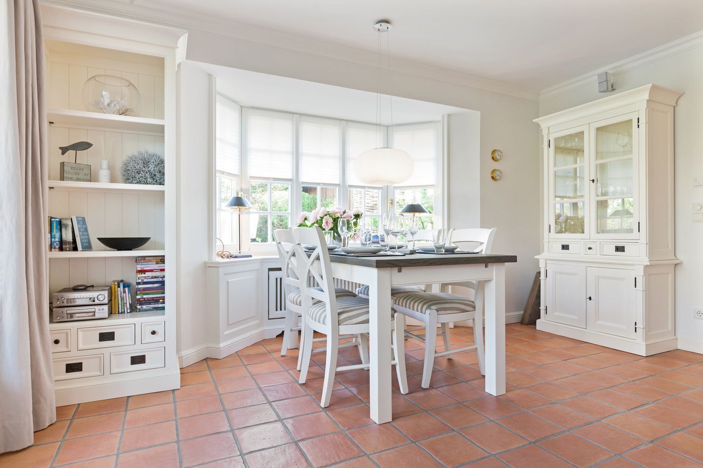 A dining area with a white table set for four, surrounded by white chairs. There is a window with a view of greenery outside, and a white cabinet with glass doors to the right.