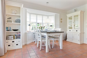 A dining area with a white table set for four, surrounded by white chairs. There is a window with a view of greenery outside, and a white cabinet with glass doors to the right.