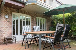 A patio with a wooden table and striped chairs, a green umbrella, and a brick wall with a window.