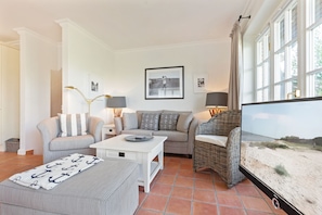 Una sala de estar con un sofá, sillones, una mesa de centro y una televisión. Hay cuadros enmarcados en la pared y una ventana con cortinas.