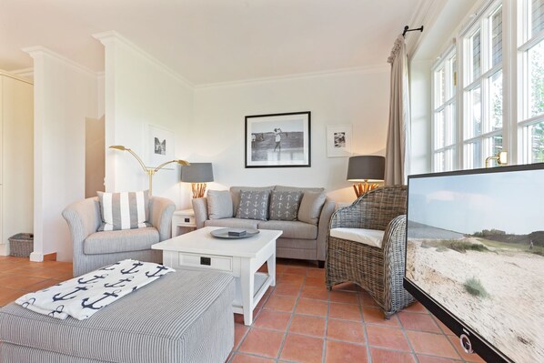 A living room with a sofa, armchairs, a coffee table, and a television. There are framed pictures on the wall and a window with curtains.