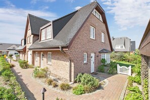 Une maison à deux étages, avec une façade en briques, un toit en pente et un petit jardin devant, agrémenté d’un chemin et de quelques plantes.