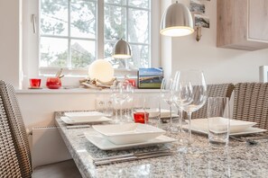 Un comedor con encimera de granito, platos blancos y copas de cristal. Hay sillas de mimbre y una lámpara colgante.