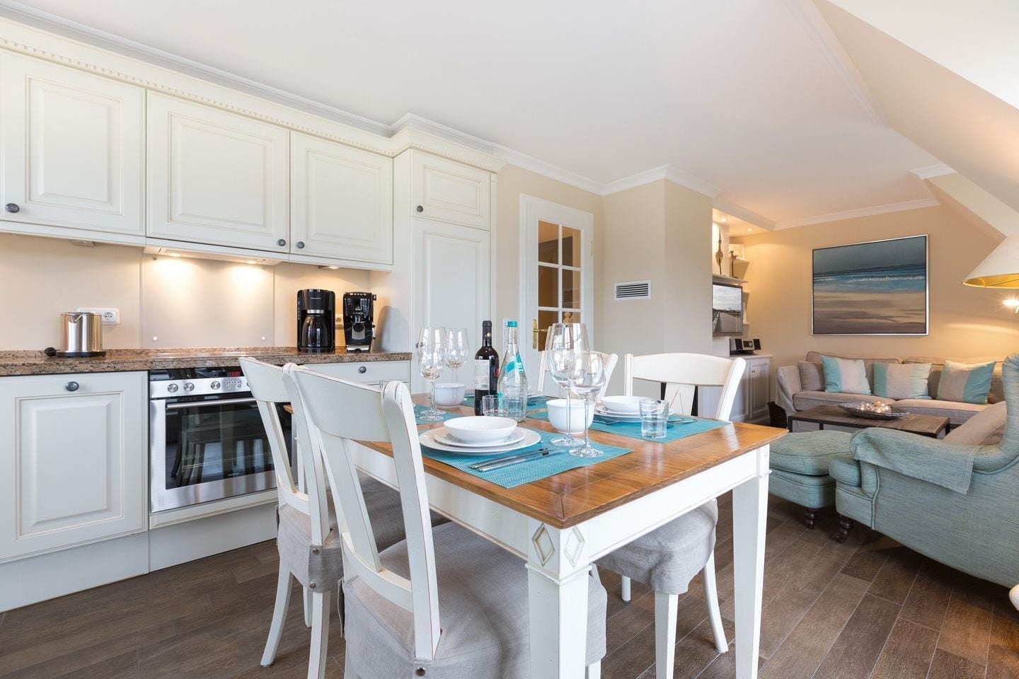 Une cuisine moderne avec des meubles blancs, un coin repas avec une table et des chaises en bois, et un salon avec un canapé et un tableau au mur.