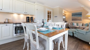 Une cuisine moderne avec des meubles blancs, un coin repas avec une table et des chaises en bois, et un salon avec un canapé et un tableau au mur.