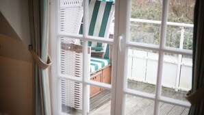 Une vue à travers une fenêtre à cadre blanc, qui révèle une terrasse avec un plancher en bois, une clôture blanche et une chaise à rayures vertes et blanches.