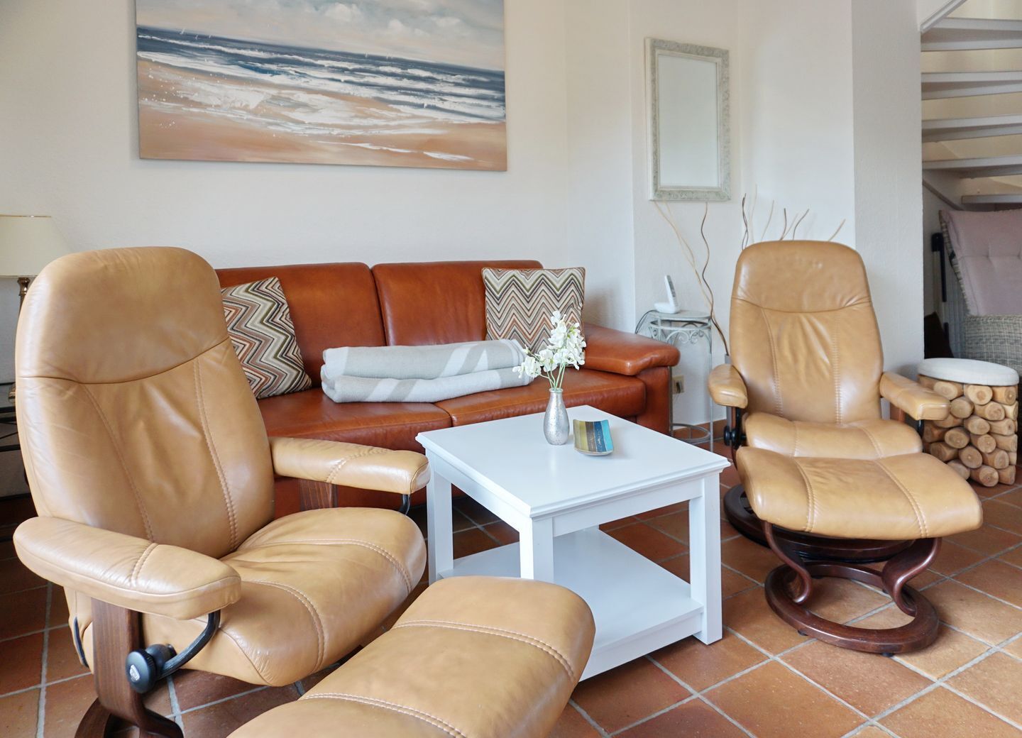 A living room with a brown leather sofa, two leather armchairs, a white coffee table, and a painting of a beach scene.
