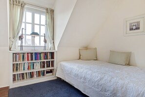 Une chambre avec un plafond en pente, une bibliothèque remplie de livres, un lit recouvert d’une couette blanche et un tableau encadré accroché au mur.