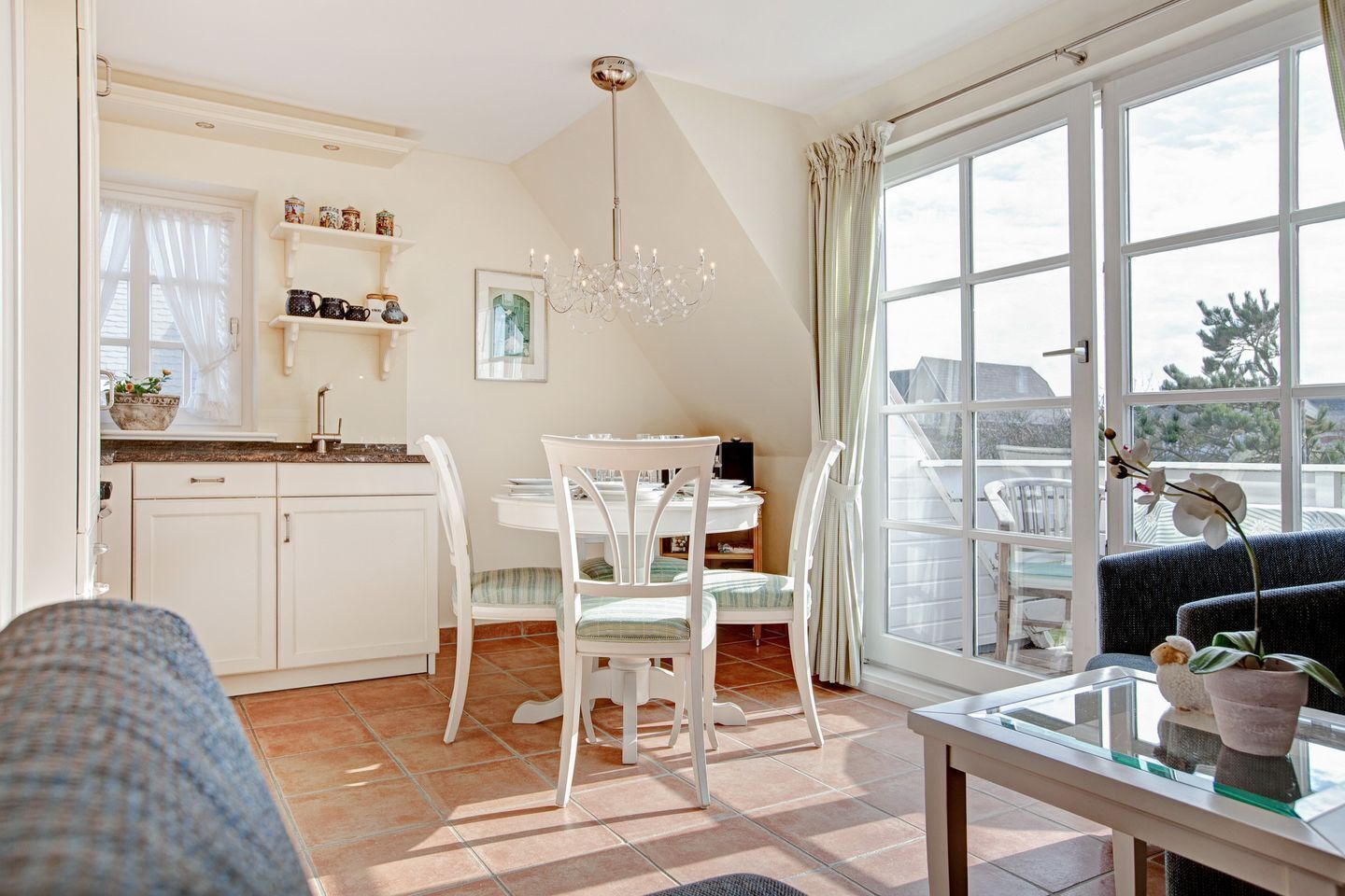A bright kitchen with white cabinets, a dining table with chairs, a chandelier, and a large window with curtains.