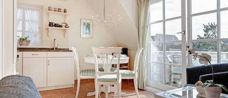 A bright kitchen with white cabinets, a dining table with chairs, a chandelier, and a large window with curtains.