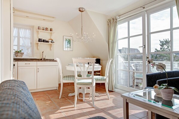 A bright kitchen with white cabinets, a dining table with chairs, a chandelier, and a large window with curtains.