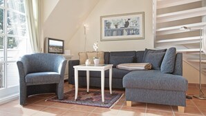 A living room with a grey sofa, a blue armchair, a small white table with a plant, and a framed picture on the wall. - Friesenhörn, App. 4 Links - Dorfteich. Dünen. Viel Erholung (Wenningstedt-Braderup (Sylt))