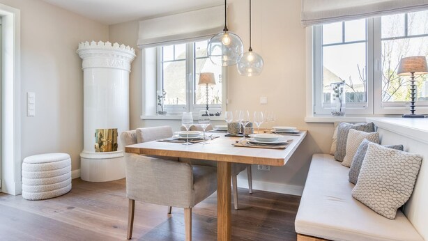 Un comedor con mesa de madera, sillas y un banco. Hay ventanas con cortinas, una araña y una lámpara.