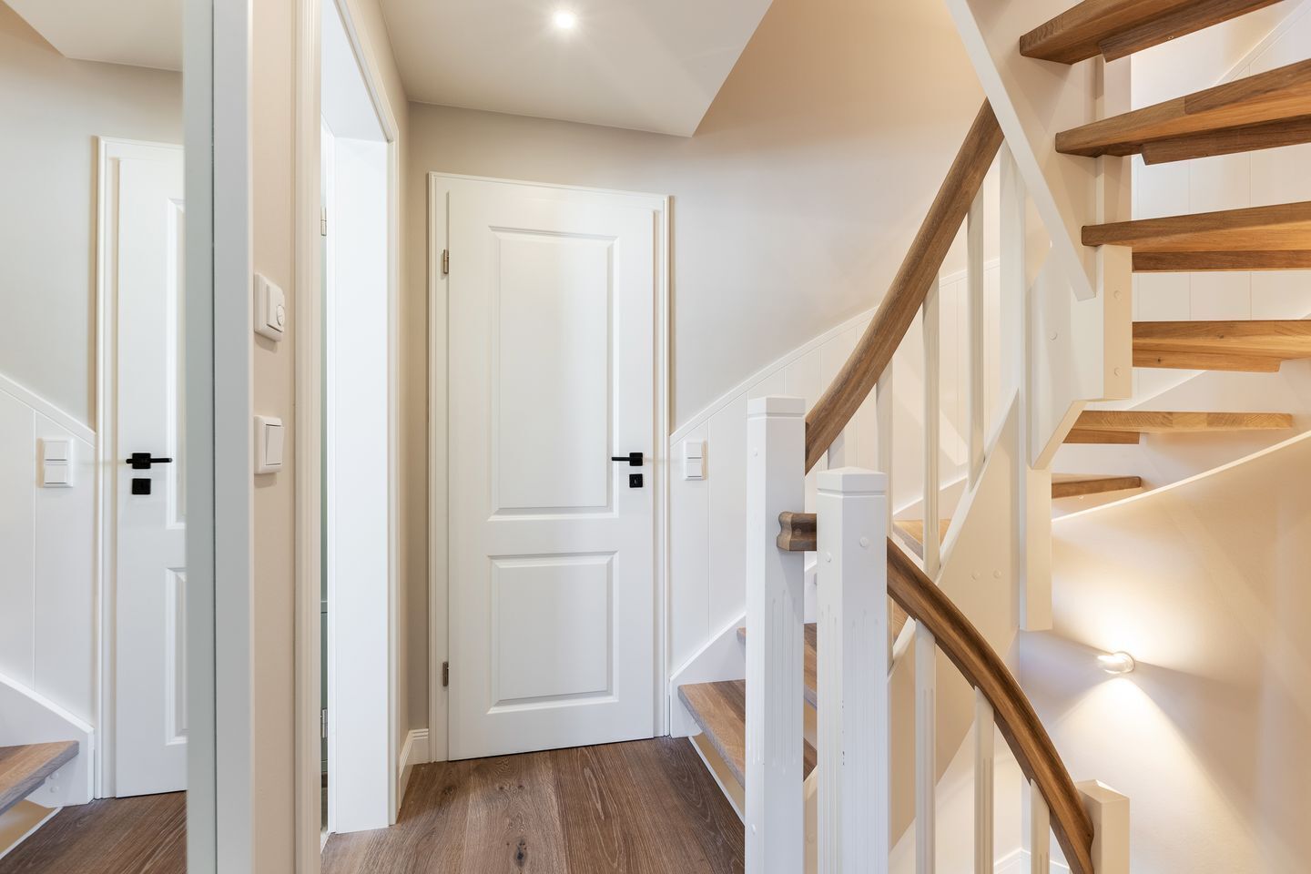 A modern interior with a wooden staircase, white walls, and a door at the end of the hallway.