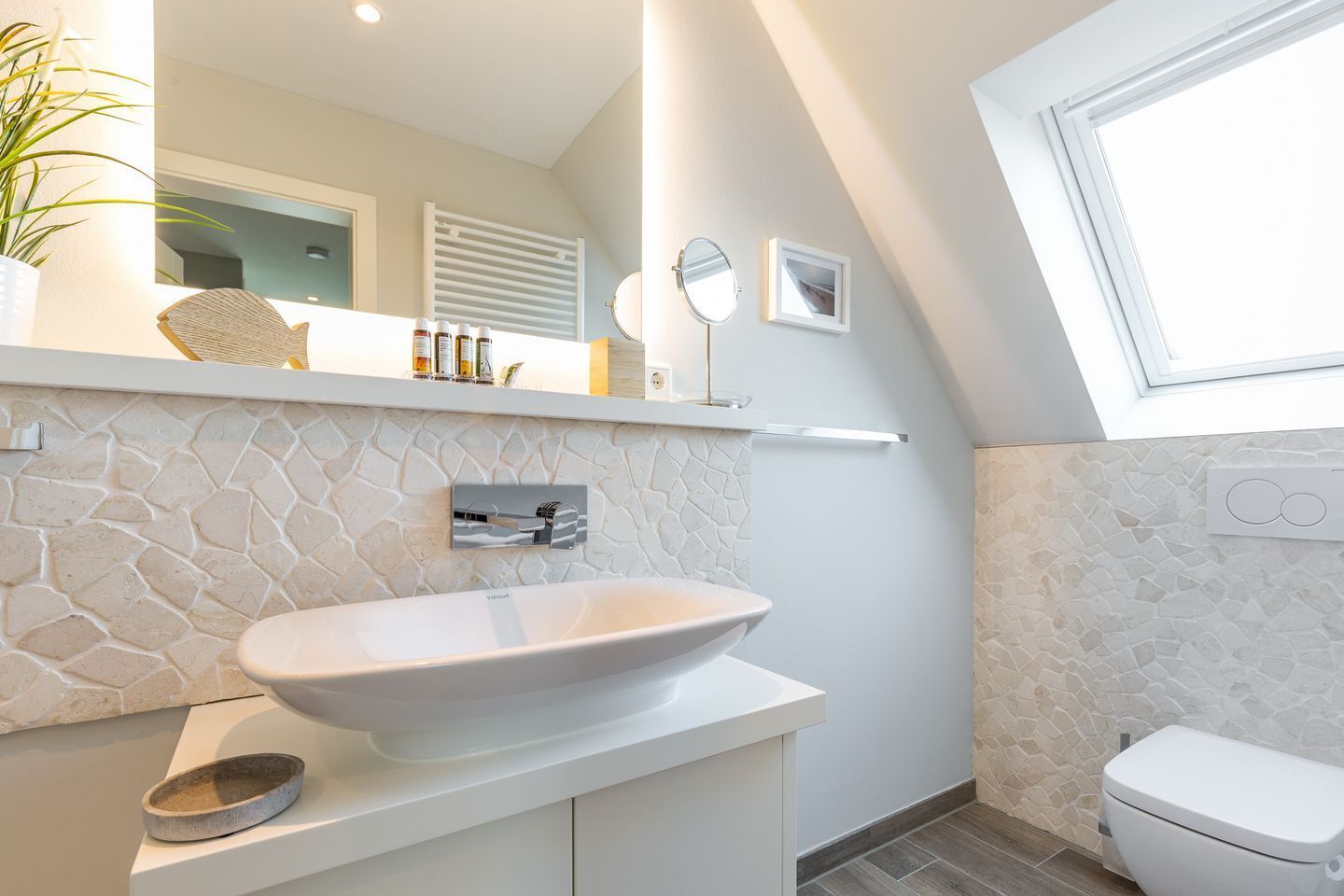 Un baño moderno con salpicadero de piedra, un lavamanos blanco, un inodoro y una ventana que proporciona luz natural.