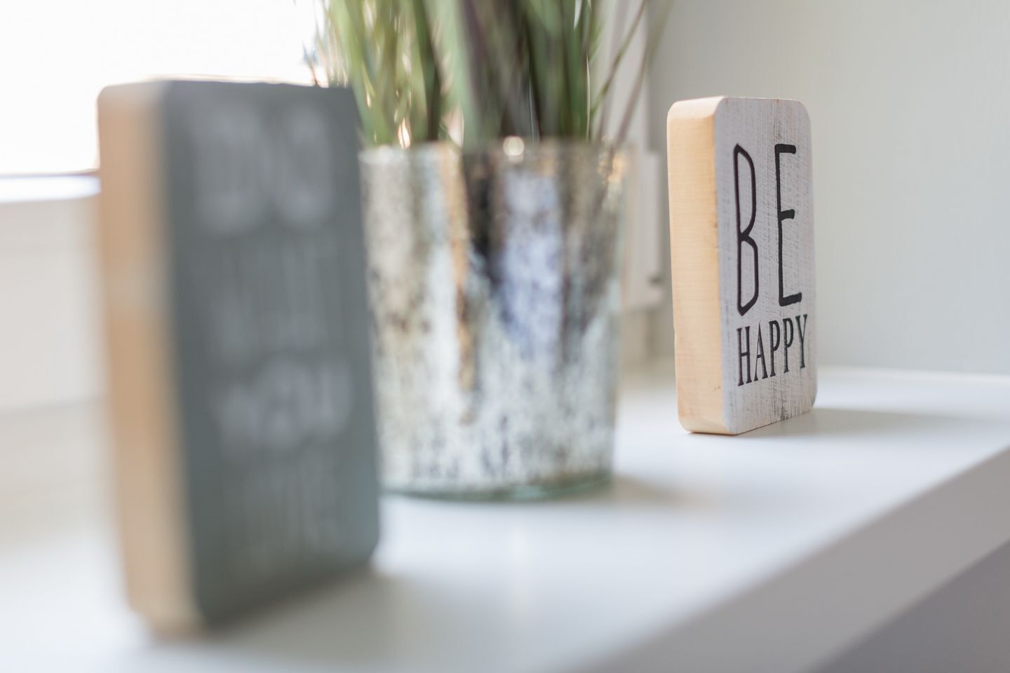 Ein hölzerner Block mit der Aufschrift 'BE HAPPY', eine Glasvase mit Pflanzen und ein verschwommenes Buch auf einer weißen Fläche.