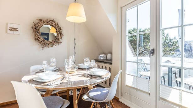 A dining area with a round table set for four, a pendant light, and a view through a large window.