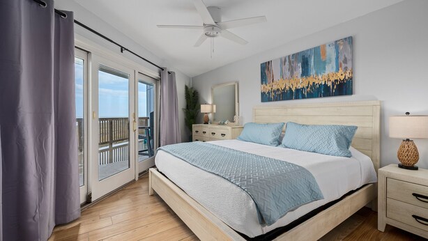 A bedroom with a large bed, a ceiling fan, a nightstand, a lamp, a painting, and a view of the outdoors through a sliding door.