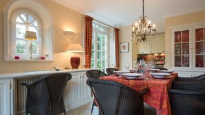 Ein Esszimmer mit Tisch für vier Personen, umgeben von Korbstühlen, einer Kristallleuchter und einem Fenster mit Vorhängen.