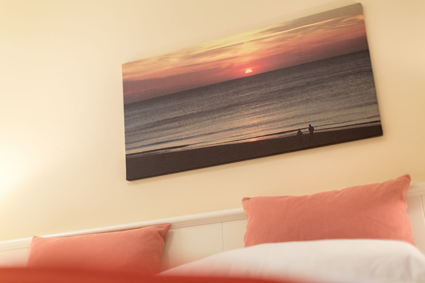 A bedroom with a wall-mounted sunset painting, a white headboard, and two pink pillows.