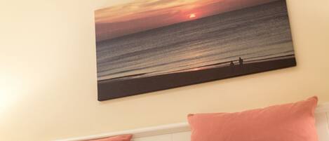 A bedroom with a wall-mounted sunset painting, a white headboard, and two pink pillows.