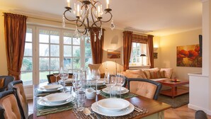 A dining area with a table set for six, surrounded by wicker chairs, a chandelier, and a view of the garden through large windows. - Klingler's Hüs, App. 1 - Style Meets Fashion. Bestlage Zwischen Dünen und Dorfteich (Wenningstedt-Braderup (Sylt))