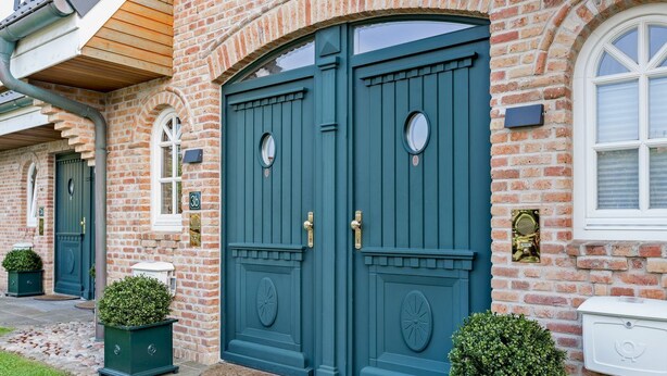 A two-story brick house with a large, dark green double door, a small window with a white frame, and a mailbox on the right side. - Klingler's Hüs, App. 1 - Style Meets Fashion. Bestlage Zwischen Dünen und Dorfteich (Wenningstedt-Braderup (Sylt))