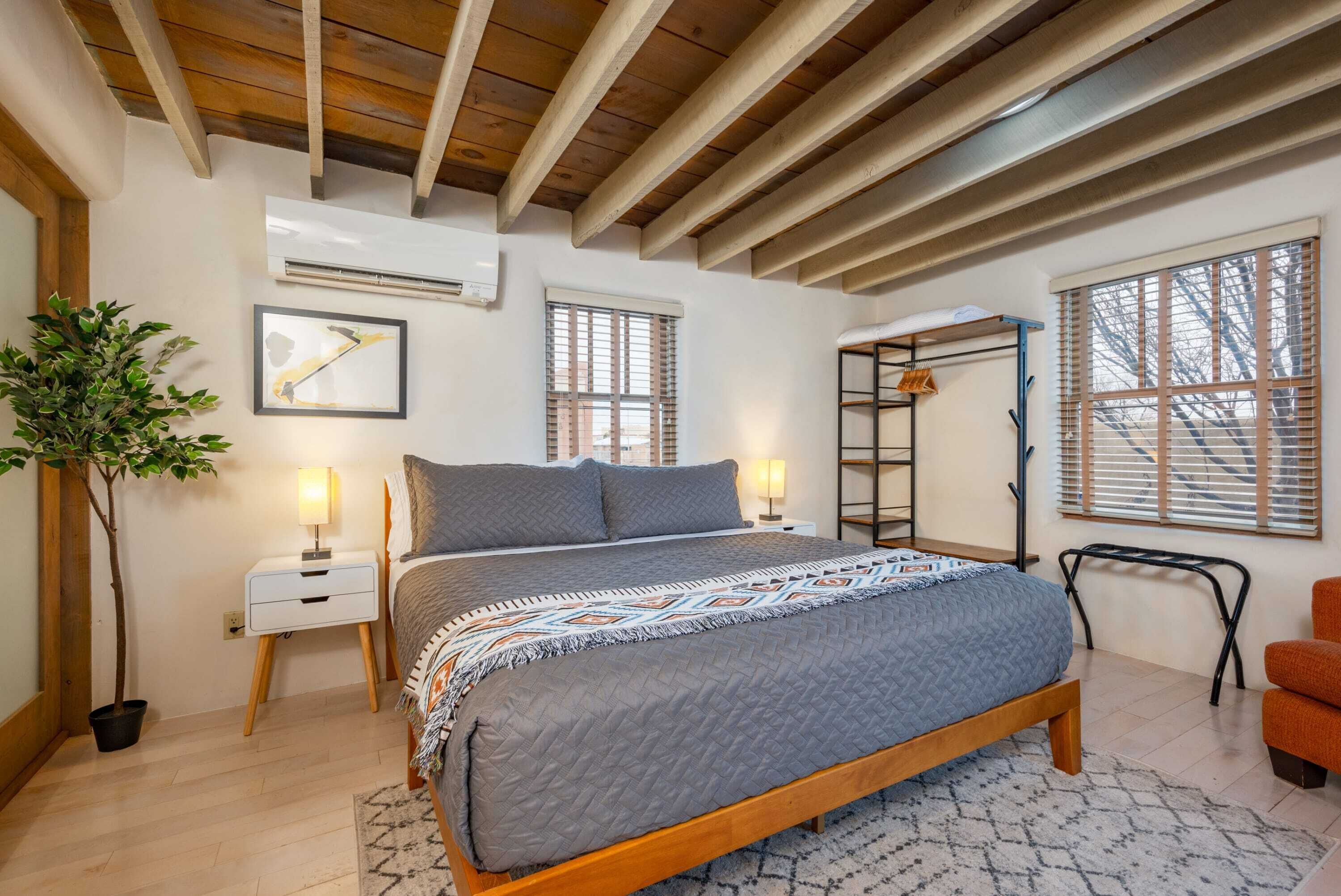 Quarto com cama de madeira, mesa de cabeceira, planta em vaso, janela com persianas e uma escada encostada na parede.