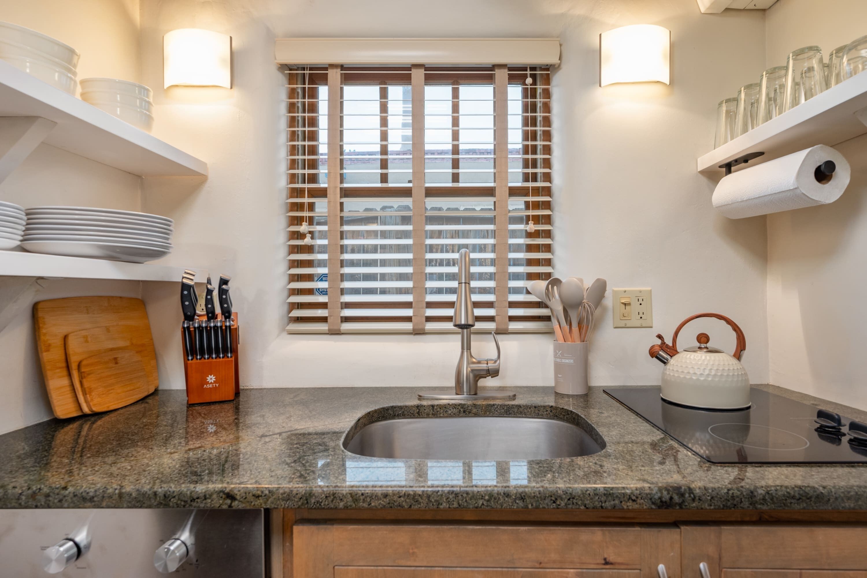 Una cocina con encimera de granito, pileta de acero inoxidable, muebles de madera, una ventana con persianas y un hervidor sobre una cocina por inducción.