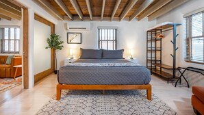 Un dormitorio con techo de madera, una cama con colcha gris, una mesita de noche con lámpara y una ventana con persianas.