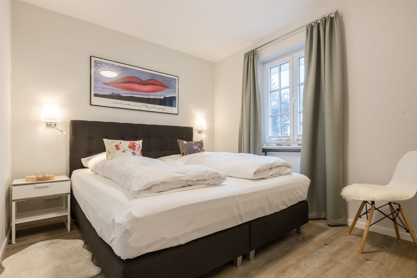 Una camera da letto con un letto, comodini, una sedia, una finestra con tende e un’opera d’arte appesa al muro.