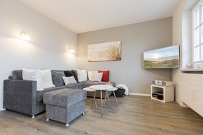A modern living room with a grey sectional sofa, a small round table, and a flat-screen TV mounted on the wall. - Haus am Meer, Strandheuler App. 2 - Hyggeliges Wohngefühl Pur! Ein Katzensprung zum Strand! Alles (Wenningstedt-Braderup (Sylt))