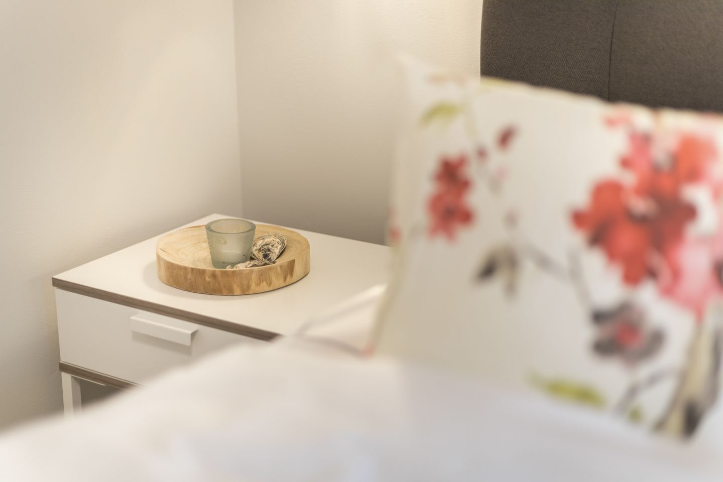 Een netjes opgemaakt bed met een bloemenkussen, een nachtkastje met een glas en een klein object, en een houten dienblad.