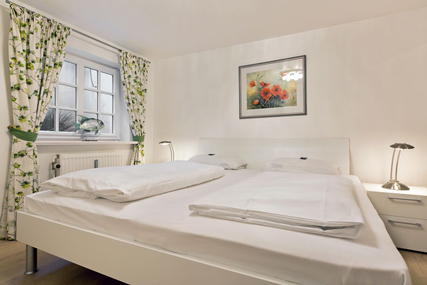 Een net opgemaakt tweepersoonsbed met witte lakens, een nachtkastje met een lamp, een radiator en een raam met gordijnen.