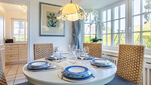A dining area with a round table set for four, surrounded by wicker chairs, a hanging light fixture, and a window with white curtains. - Meeresbrise, App. 1 - Feines Wohnen mit Viel Platz Unter Kuscheligem Reet in Bestlage (Wenningstedt-Braderup (Sylt))