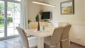 A dining area with a white table, wicker chairs, a bottle of wine, and a glass. A TV is mounted on the wall, and a painting is visible.