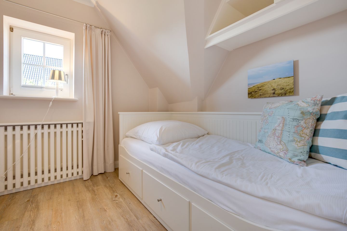 A small, neatly arranged bedroom with a single bed, a window with curtains, a radiator, and a wall-mounted map.