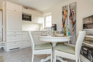 Ein Essbereich mit einem runden Tisch, gedeckt für vier Personen, umgeben von Korbstühlen mit grünen Kissen. Weiße Schränke mit einer Mikrowelle und einem Backofen über der Arbeitsplatte sind sichtbar. Zwei abstrakte Gemälde hängen an der Wand.