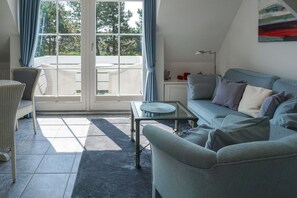 Un salon avec un canapé, un fauteuil et une petite table. Il y a une grande fenêtre avec des rideaux, une lampe et un tableau au mur.