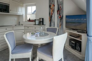 A compact kitchen with white cabinetry, a dining area with wicker chairs, a coffee maker, and a flat-screen TV on a stand.