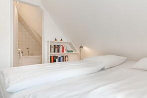 Une chambre avec un lit blanc, une bibliothèque et une salle de bain avec douche.