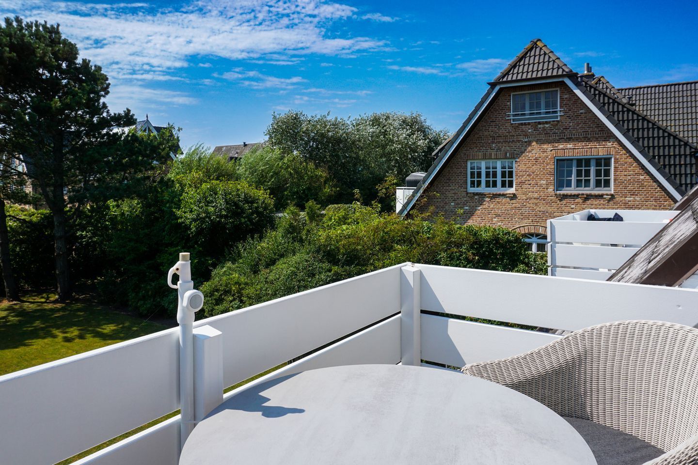 A rooftop terrace with a white railing, a round table, and a wicker chair. In the background, there is a brick house with a pitched roof and a window.