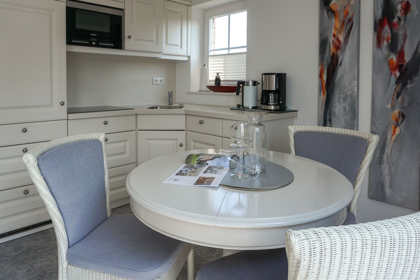 Une cuisine compacte avec des meubles blancs, un four à micro-ondes, une machine à café et un coin repas comprenant des chaises en osier et une table ronde.