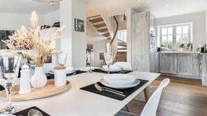 A modern dining area with a wooden table set for four, white chairs, and a vase with dried pampas grass. A staircase leads to an upper level, and a kitchen with a wooden countertop is visible in the background.