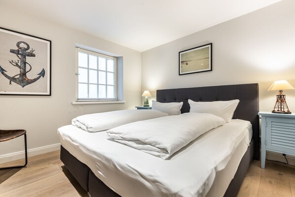 Cama doble con ropa blanca, mesita de noche con lámpara, ventana con persianas y obras de arte en las paredes.