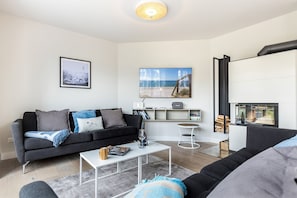 Ett modernt vardagsrum med en svart soffa, ett soffbord och en tv. Det finns en bokhylla med böcker och ett litet bord med en stol.