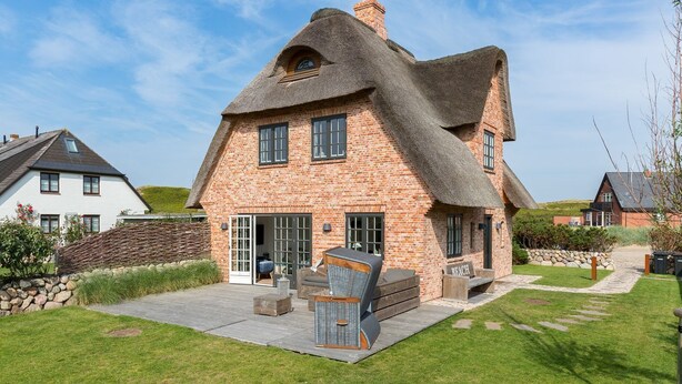 A thatched-roof house with a brick exterior, a patio with outdoor furniture, and a stone wall.