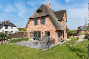 Una casa con techo de paja y fachada de ladrillo, un patio con mobiliario exterior y un muro de piedra.