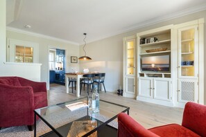 Ett modernt vardagsrum med ett soffbord i glas, röda soffor, en matplats med ett bord och stolar i trä samt ett kök med vita skåp och en tv.